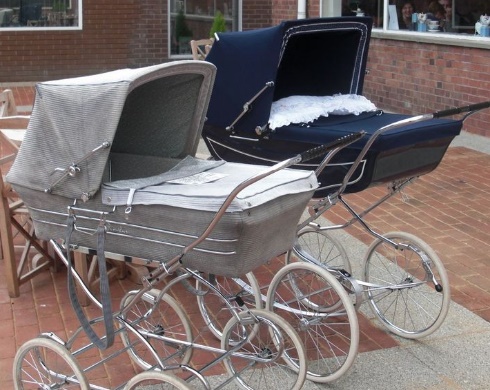 Na zdjęciu widoczne są dwa eleganckie, vintage wózki dziecięce, stylizowane na modele z dawnych czasów. Wózki mają metalowe ramy, duże, białe koła i materiałowe budki – jeden w odcieniu szaro-beżowym, drugi w granatowym. Stoją na brukowanej powierzchni, prawdopodobnie na zewnątrz kawiarni lub sklepu, co podkreśla ich klasyczny urok.